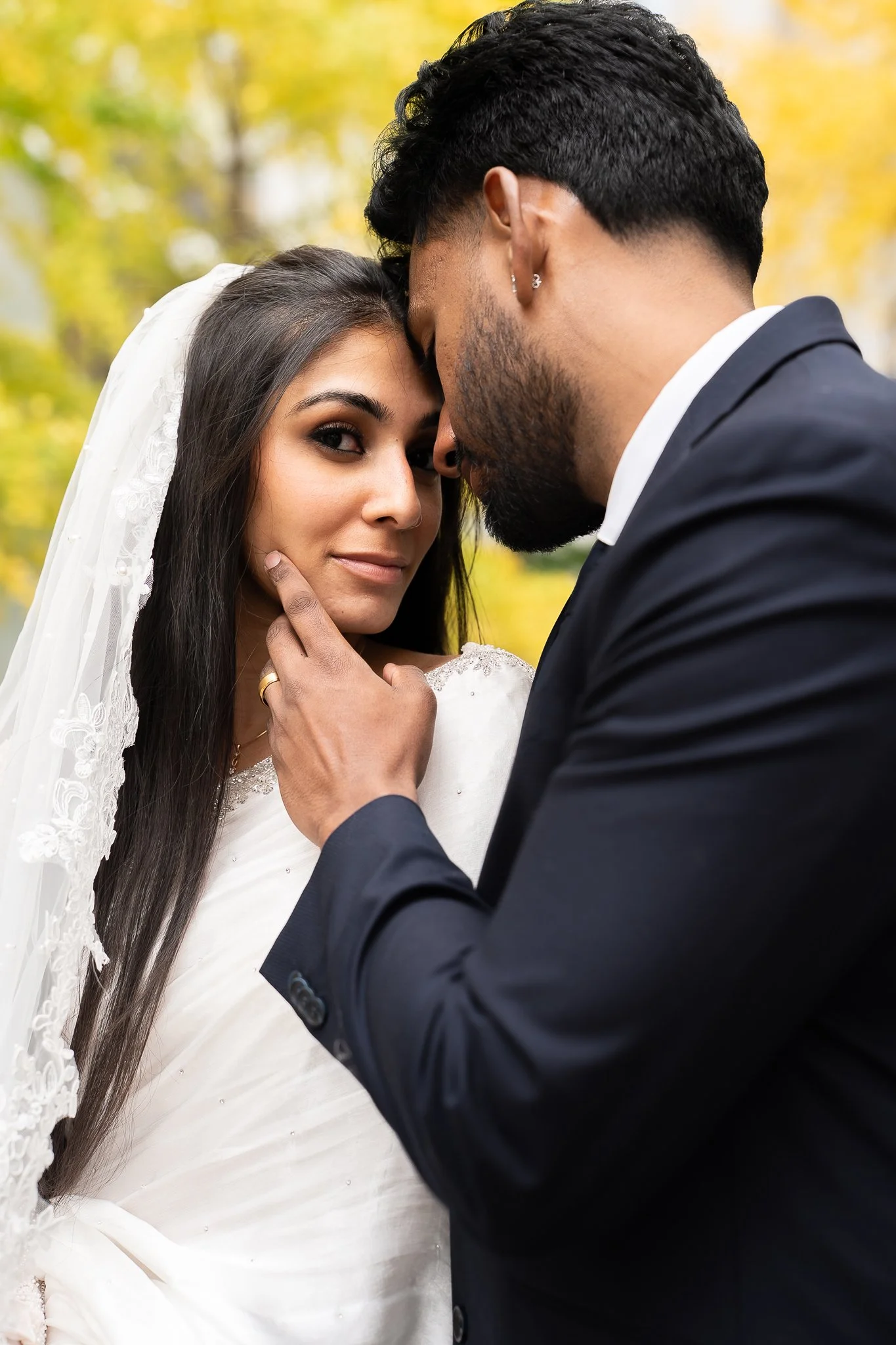 A man and woman in wedding attire close together outdoors, with the man gently touching the woman's face while they look into the camera, surrounded by autumn leaves.