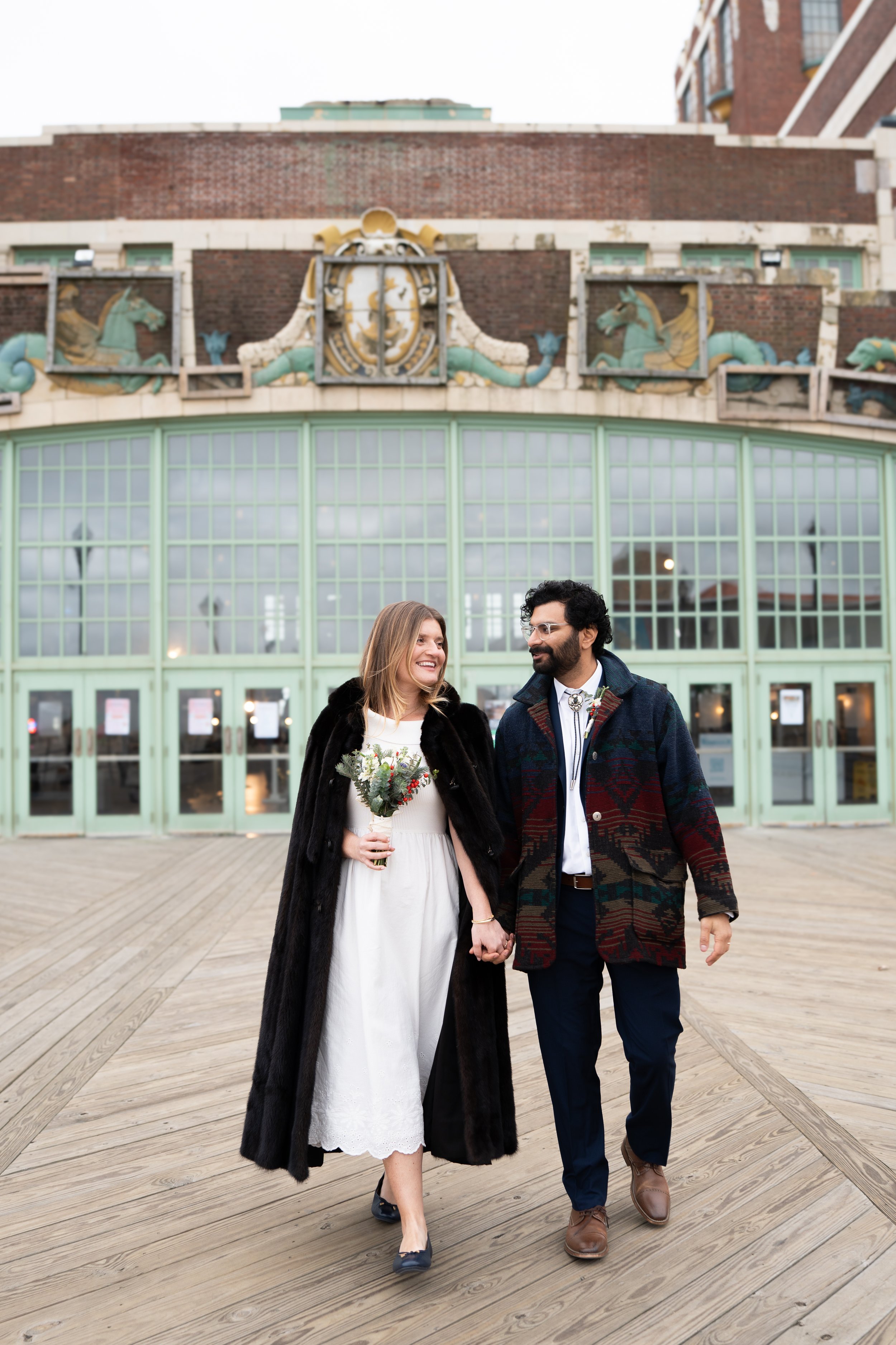 A smiling woman in a white dress holding a bouquet, and a man in a patterned coat holding her hand, walking on a wooden boardwalk in front of a building with large glass windows and decorative dragon and clock sculptures on top.
