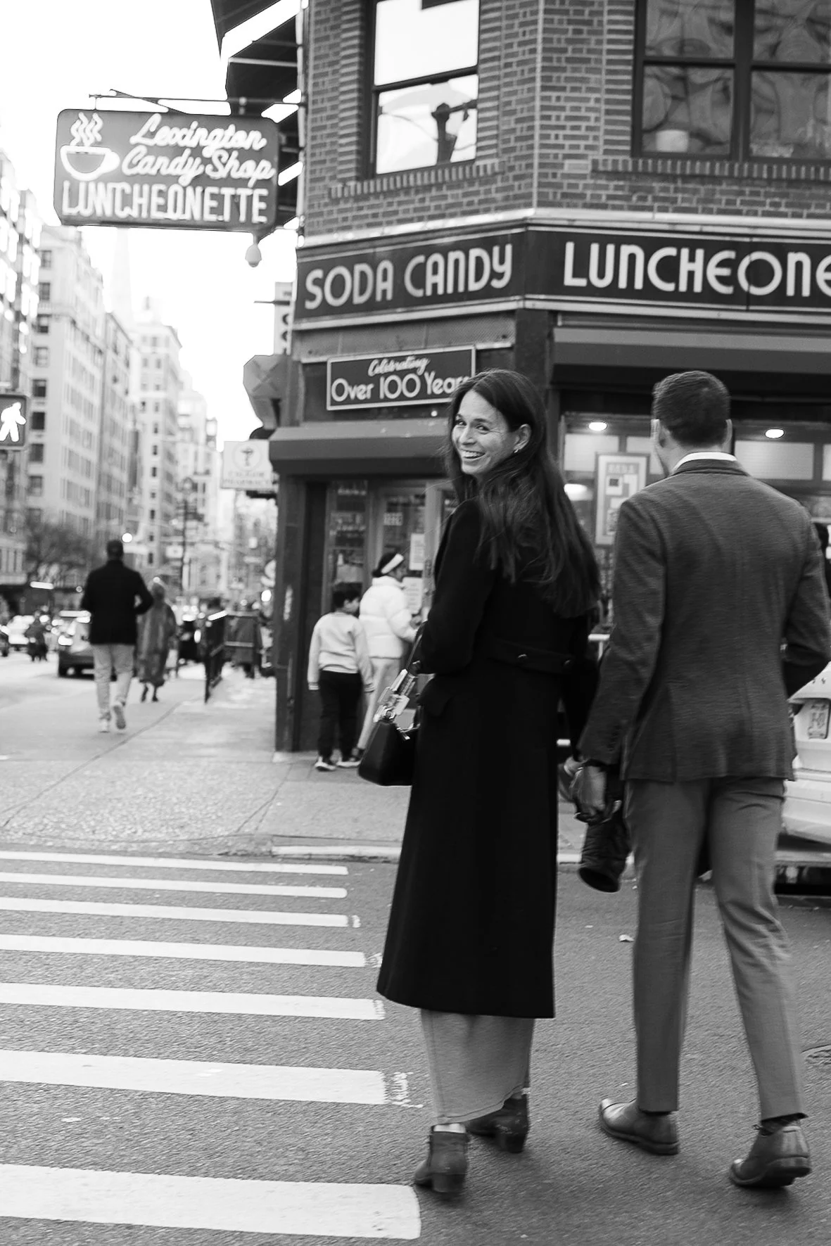 A woman crossing the street and looking back while smiling, with a man in a suit next to her, in front of a building with signs for a candy shop and luncheonette in an urban area.