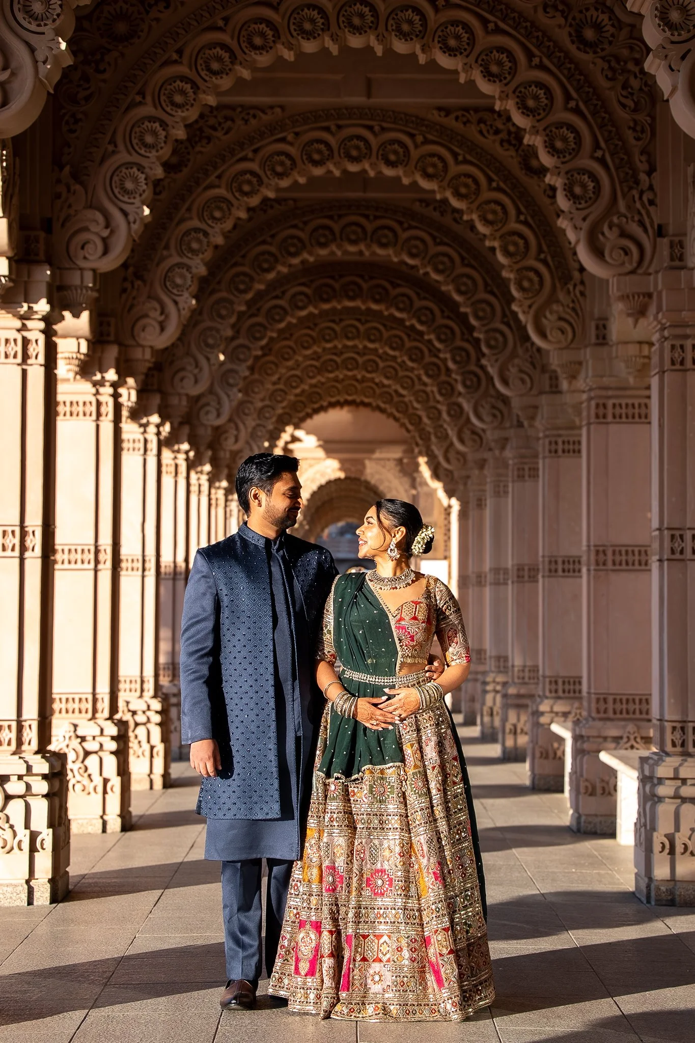 A couple dressed in traditional Indian attire stands close together, smiling at each other, in a beautifully ornate architectural corridor with arches.