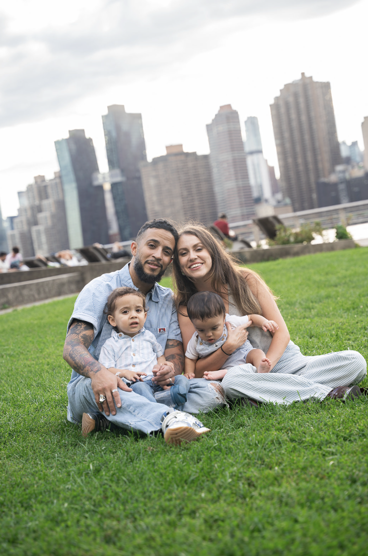 A happy family of four sitting on grass in a park with city skyscrapers in the background. The group includes a smiling man, a woman, and two young children.