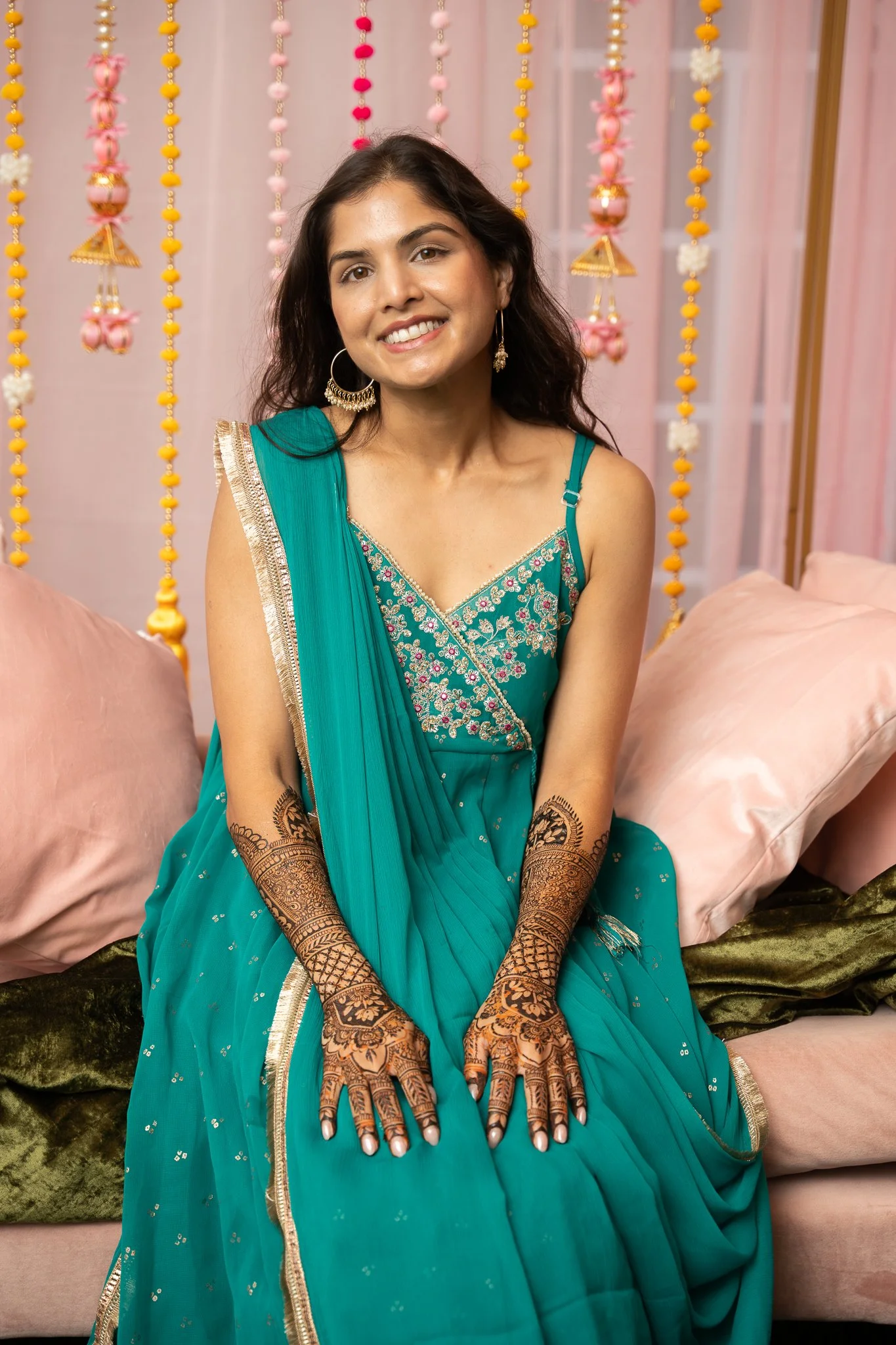 Woman sitting on a pink couch, wearing a teal sari with embroidery, gold jewelry, and mehndi designs on her arms, in a decorated room with hanging marigold and pink floral garlands.