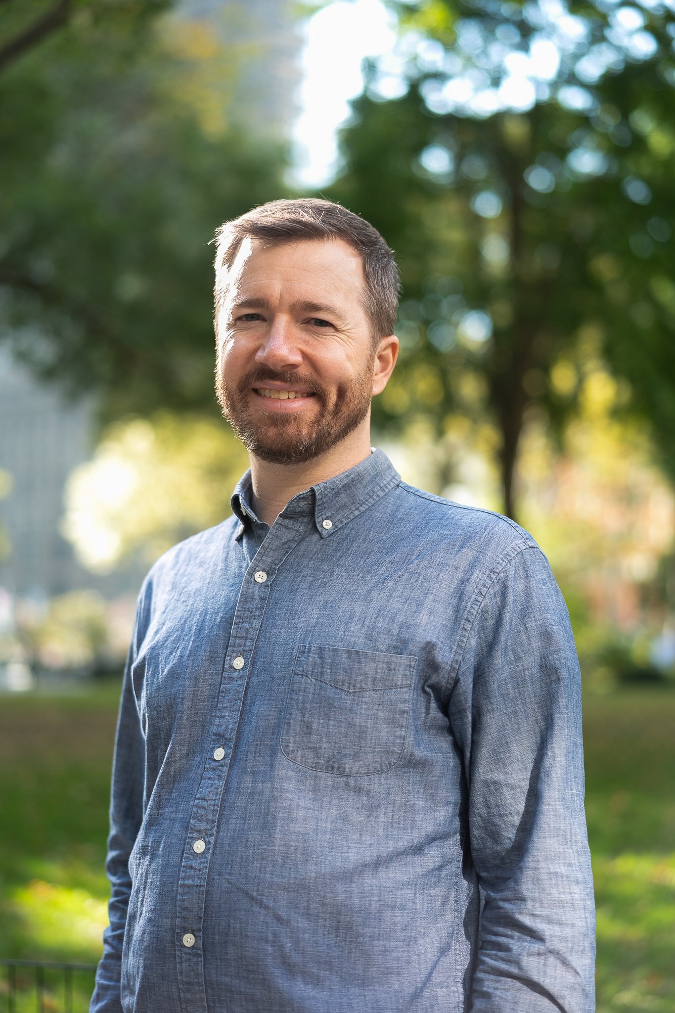 A smiling man with a beard and short brown hair, wearing a blue button-up shirt, standing outdoors in a park with green trees and sunlight in the background.