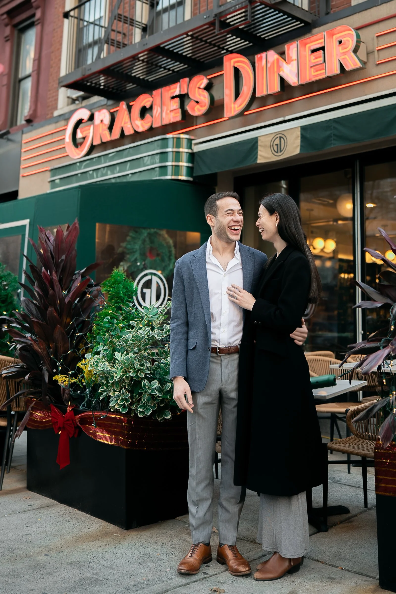 A smiling man and woman sharing a moment outside Gracie's Diner, with outdoor seating and festive plants in planters.