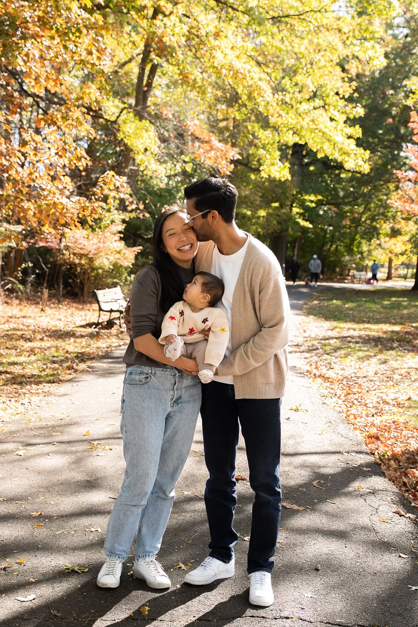A family of three standing on a park path surrounded by colorful autumn trees. The woman and man are smiling, and the man is kissing the woman's forehead. The woman is holding a young child.