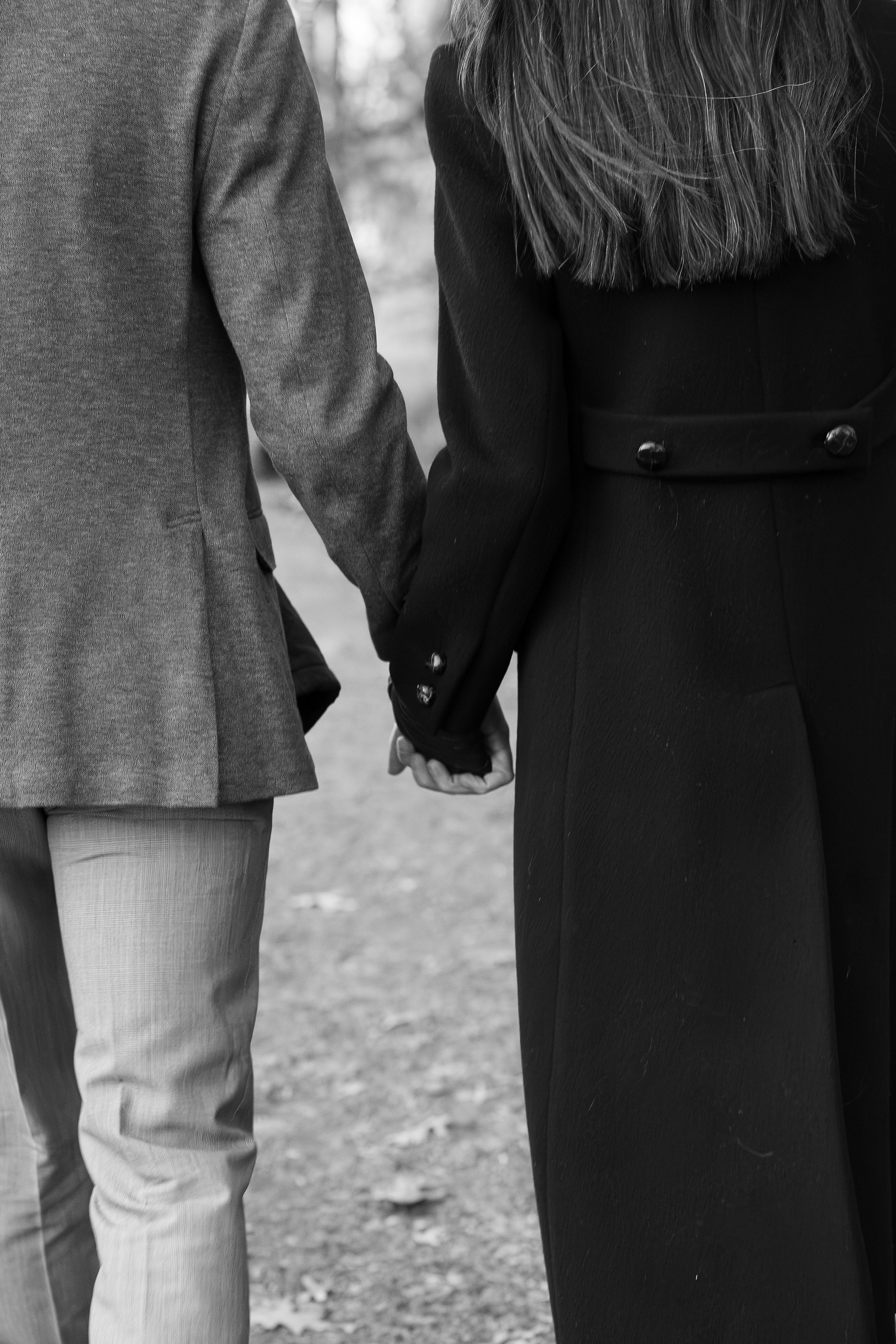 A black and white photo of a couple holding hands, with the woman wearing a dark coat and the man in a lighter suit jacket.