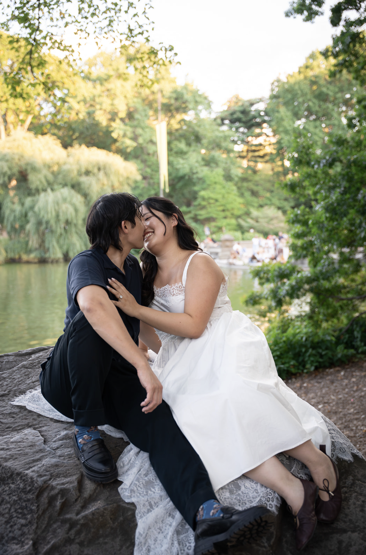 A couple sitting on a rock by a pond, affectionately kissing, surrounded by trees in a park during daylight.