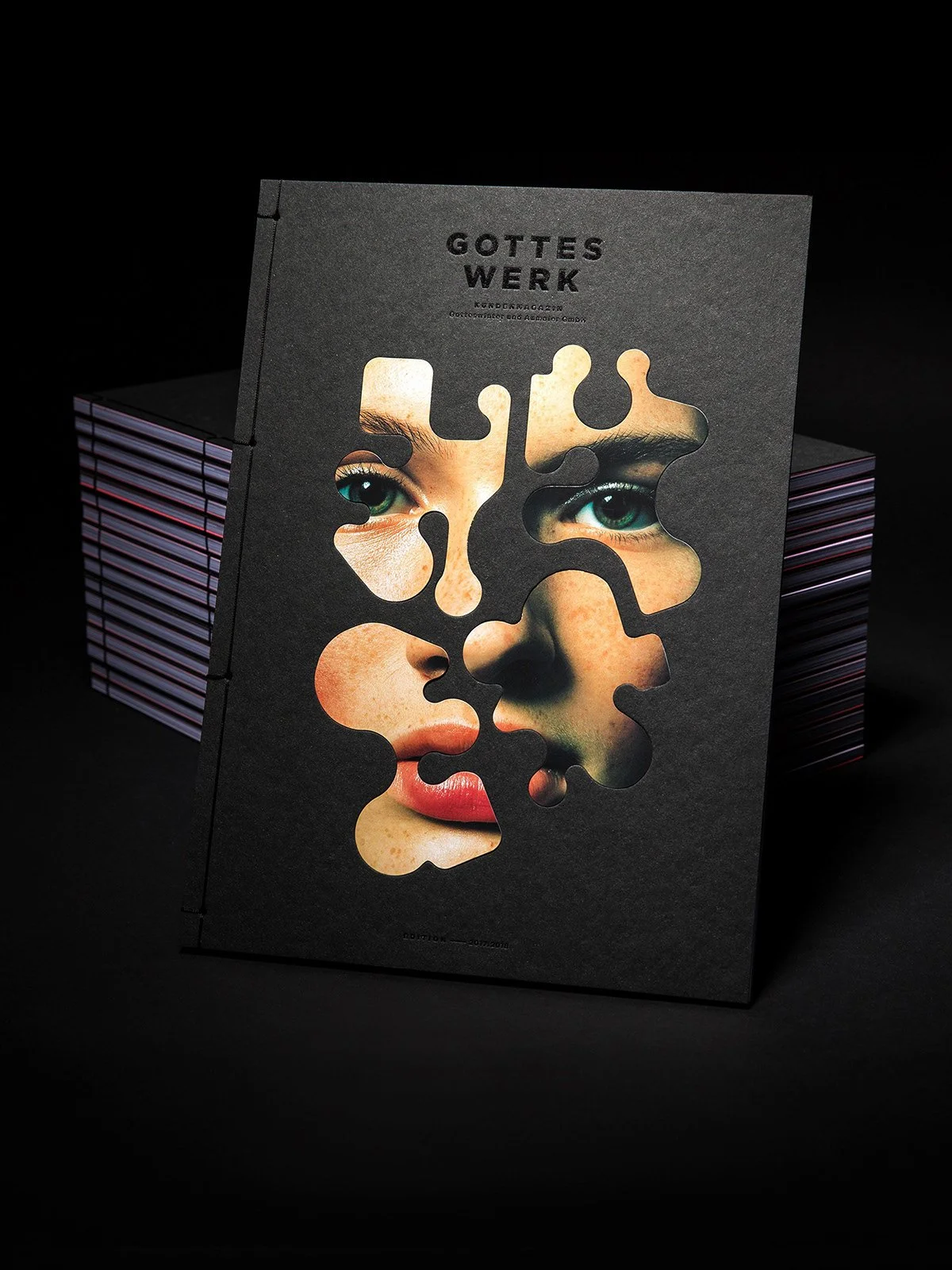 Close-up of an artistic book cover with cut-out pattern revealing a face. The book titled "Gottes Werk" is displayed with a black background and additional stacked books behind.