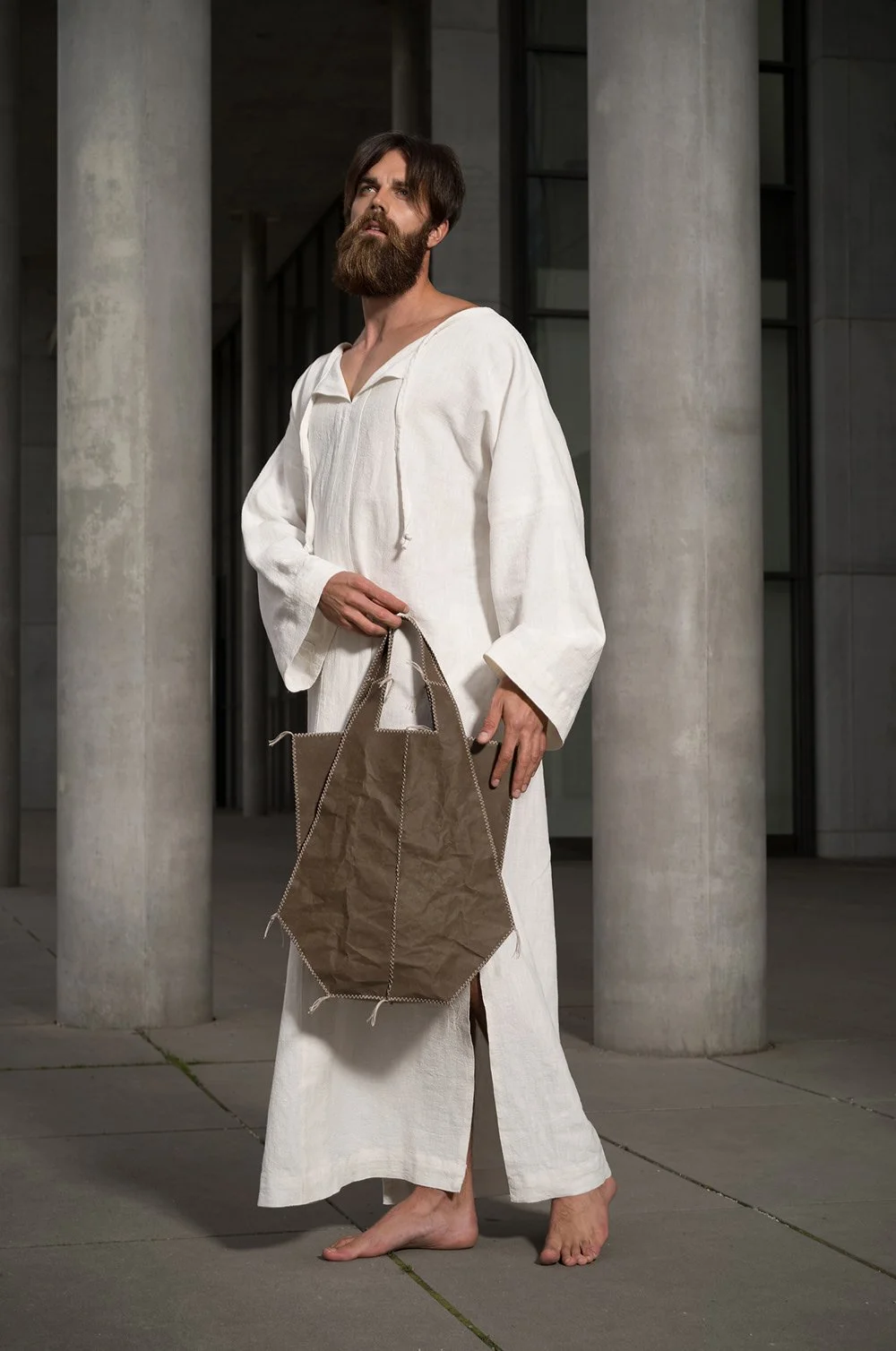 Bearded man in a white robe holding a brown bag standing barefoot next to concrete columns.