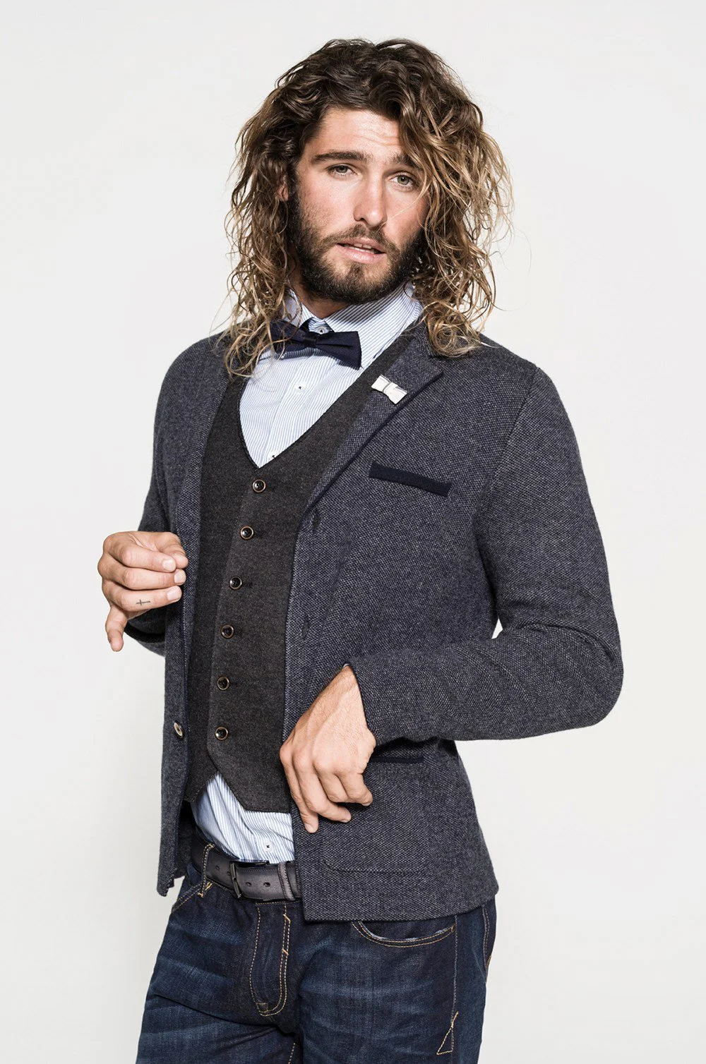 Man with long curly hair wearing a gray blazer, blue shirt, bow tie, and jeans, posing against a plain background.