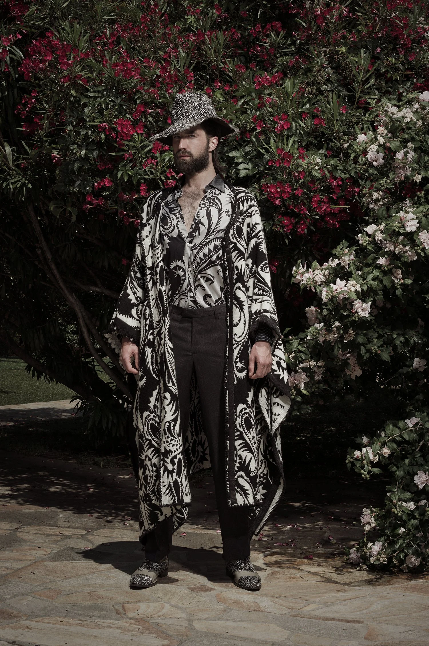 Person in elaborate black and white patterned outfit with a hat, standing in front of a bush with red and pink flowers.