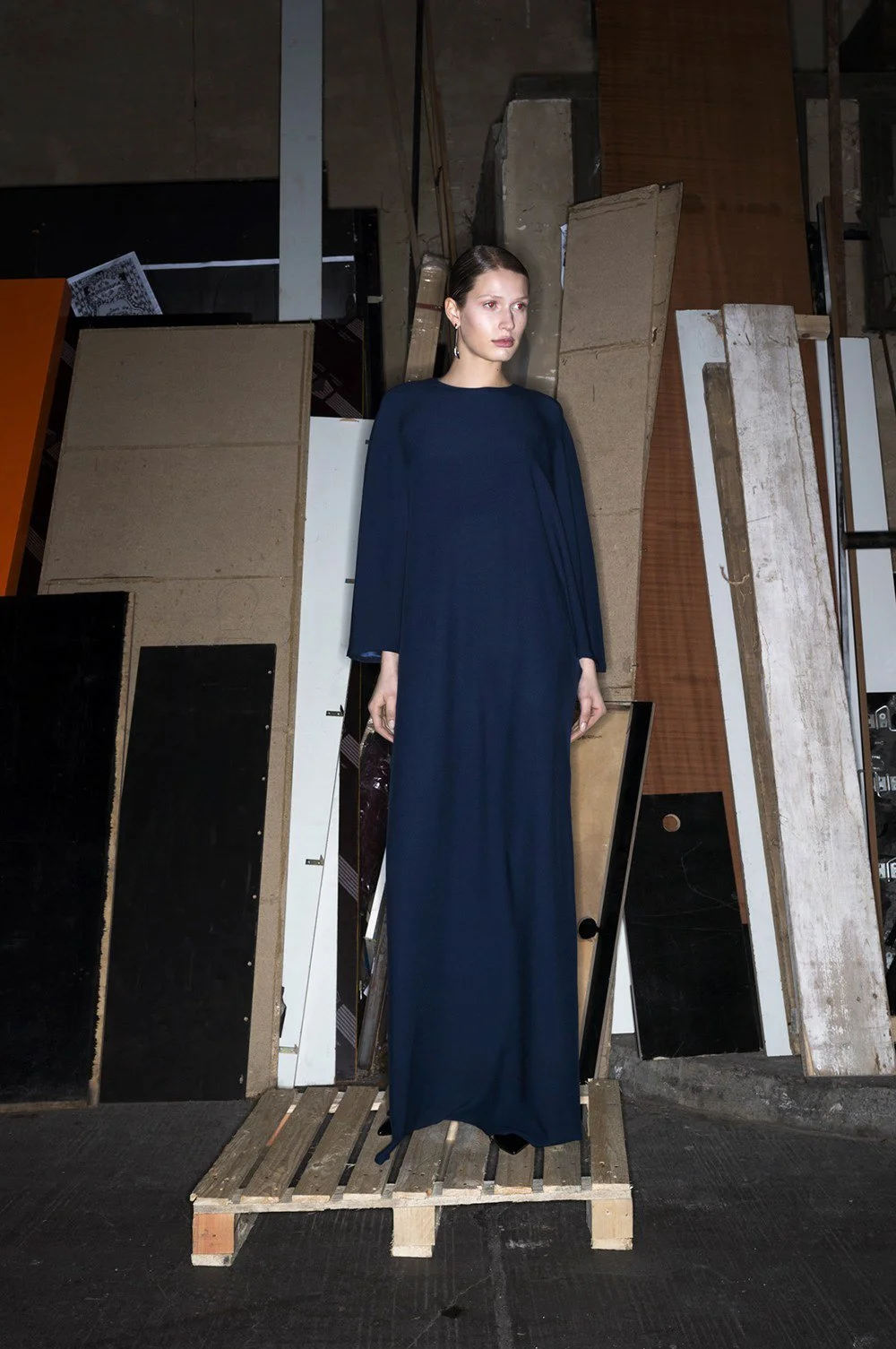 Woman in dark blue dress standing on wooden pallet with various panels in background.