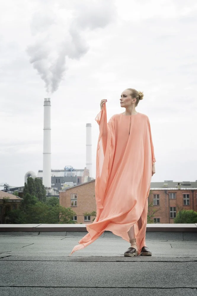 Person wearing a flowing peach dress standing on a rooftop with industrial chimneys emitting smoke in the background.