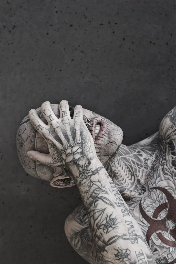 Heavily tattooed person covering their face with hand, showcasing detailed ink work on skin.