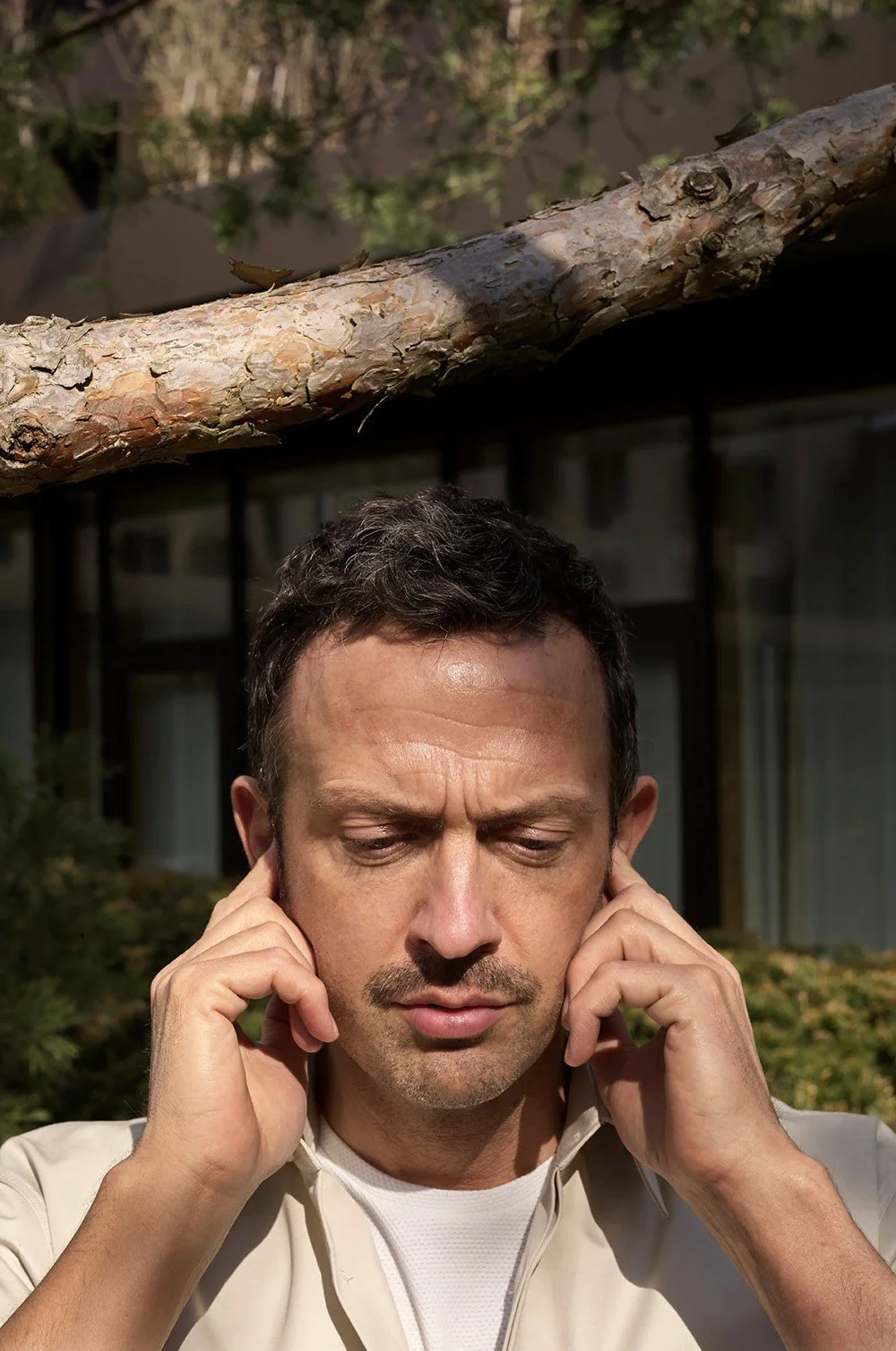 Man outdoors covering his ears with hands, with a tree branch above and a building in the background.