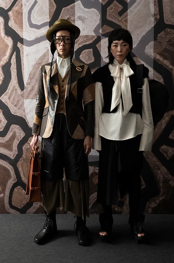Two individuals wearing eclectic fashion stand against a patterned wall. The person on the left wears a patchwork blazer, wide-brimmed hat, and glasses, holding a brown bag. The person on the right wears a white blouse with an oversized bow and dark pants, with a serious expression.