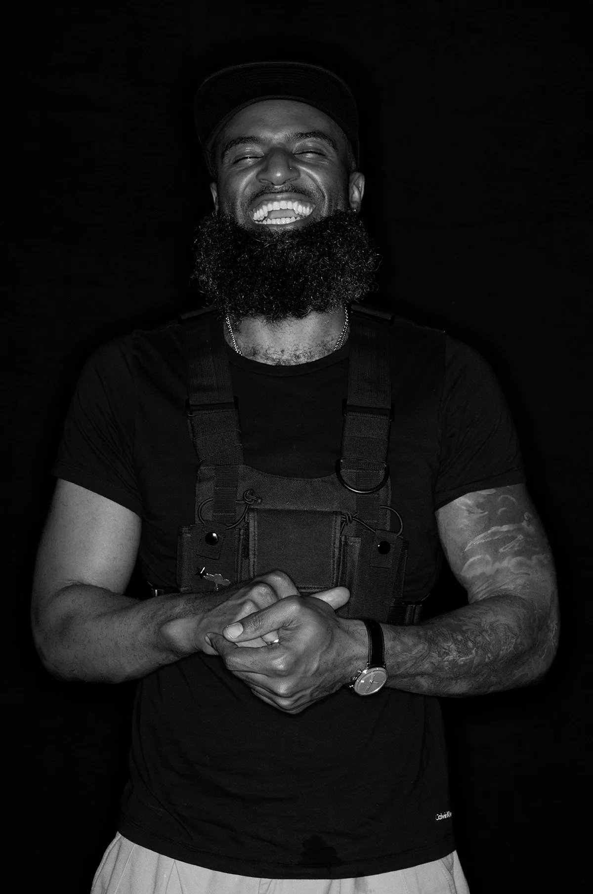 Black and white photo of a man smiling, wearing a cap, tactical vest, and watch. He has a beard and tattoos on his arms.