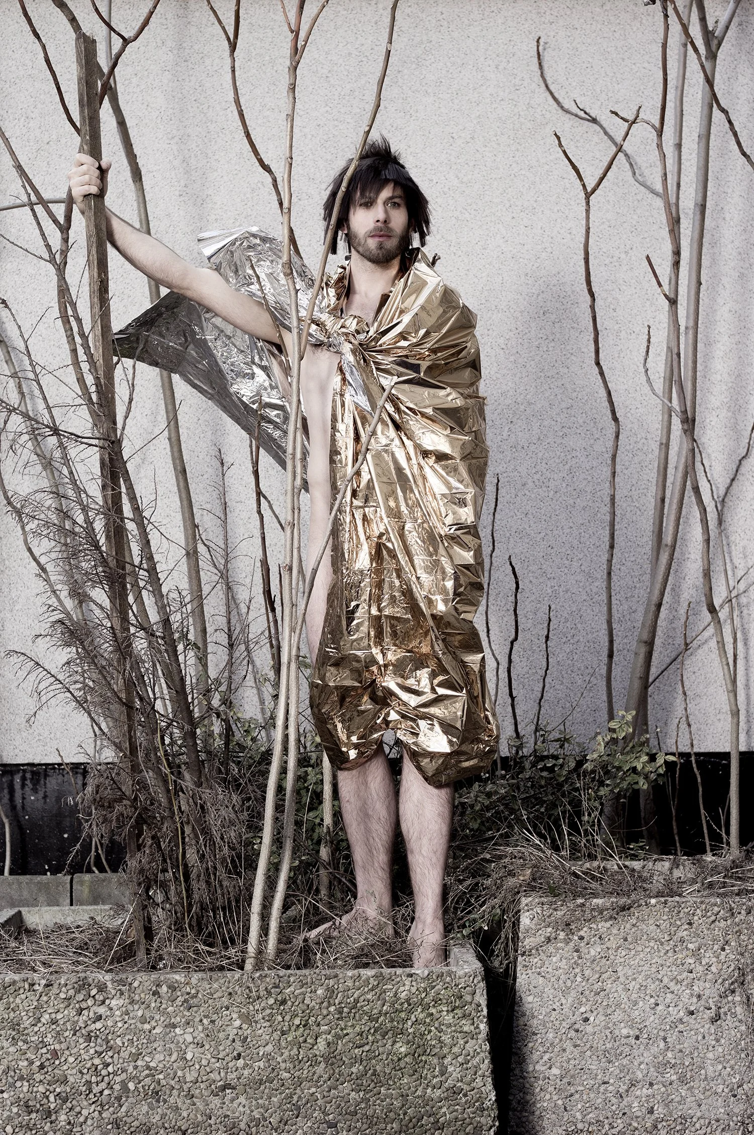 Person wrapped in metallic foil standing among concrete blocks and bare trees.