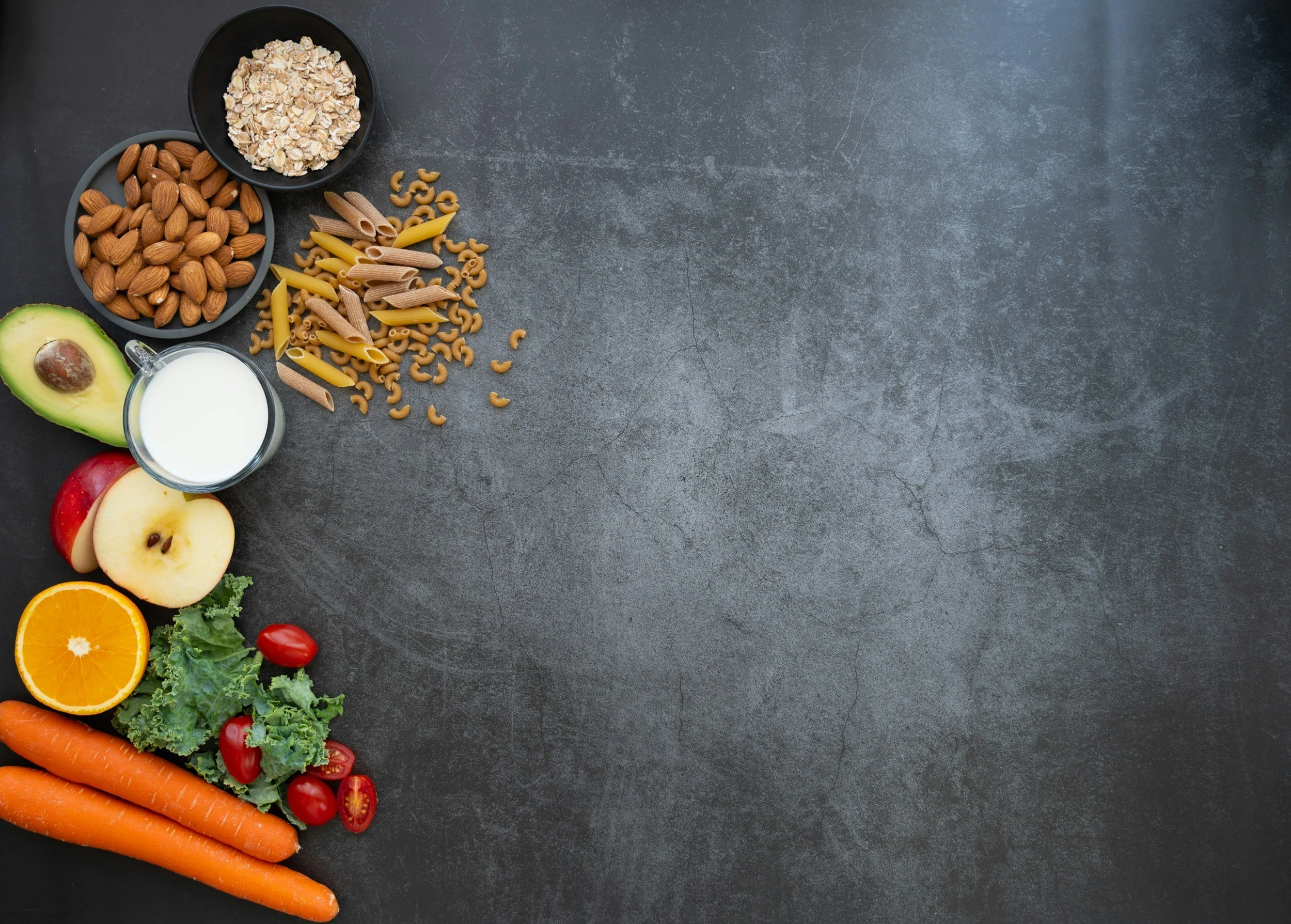 Various fresh fruits, vegetables, oats, nuts, and pasta arranged along the left side of a dark textured surface, leaving space on the right.