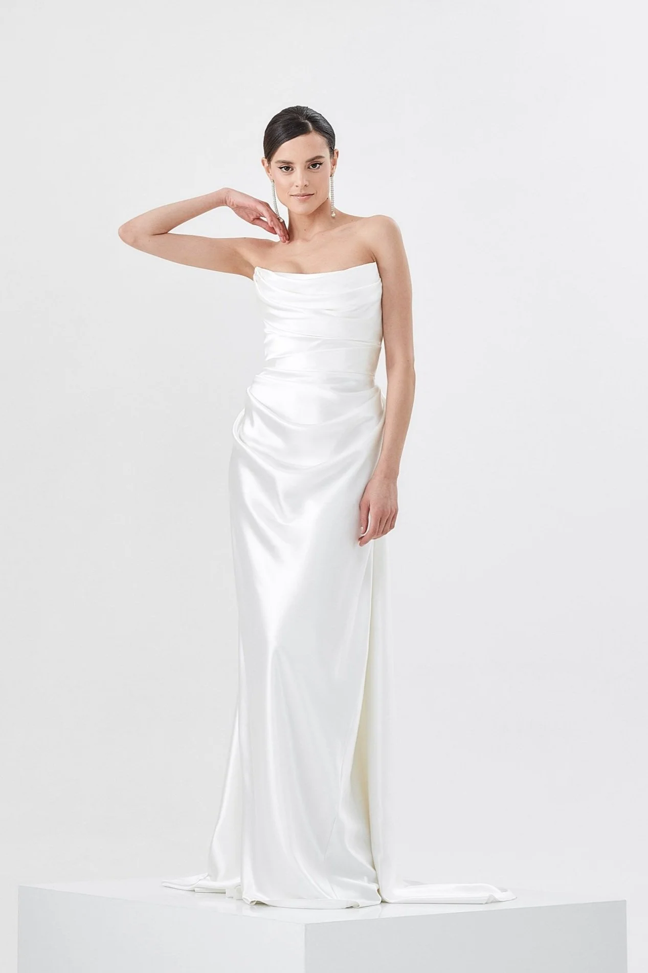 Woman in a strapless white satin gown, standing against a plain white background, with one hand near her neck and the other hanging by her side.