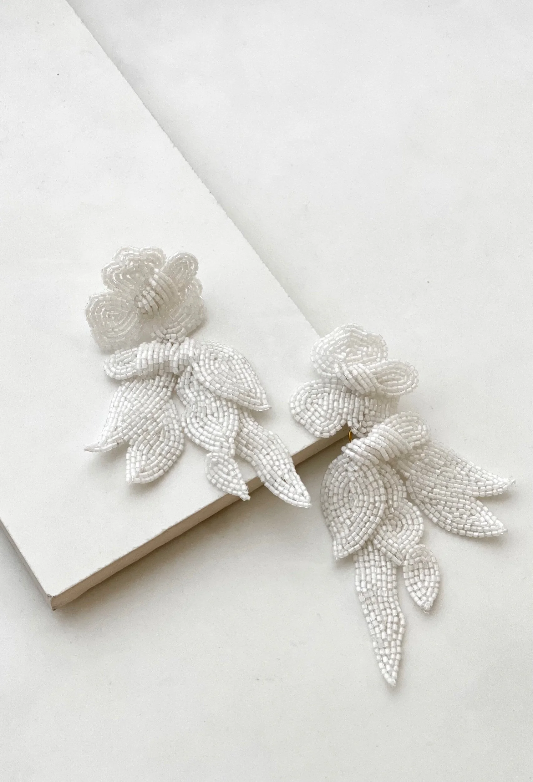 Pair of white beaded flower and leaf earrings on a white surface next to a white box.