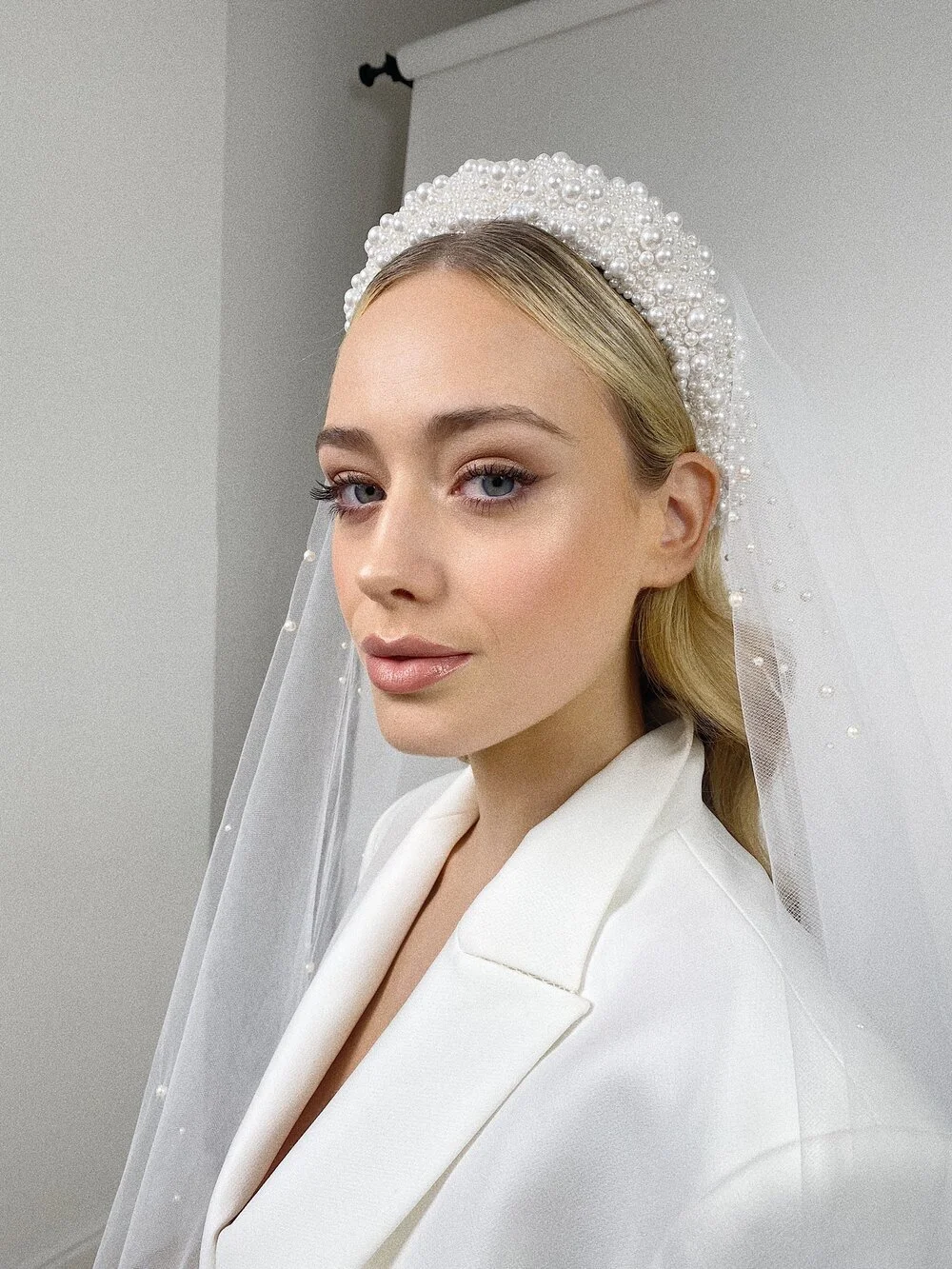 A woman dressed as a bride, wearing a pearl-studded headband, veil, and white blazer, with makeup and blonde hair styled in loose waves.
