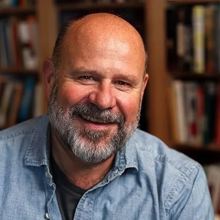 A smiling, bald man with a beard, wearing a denim shirt, in front of a bookshelf filled with books.