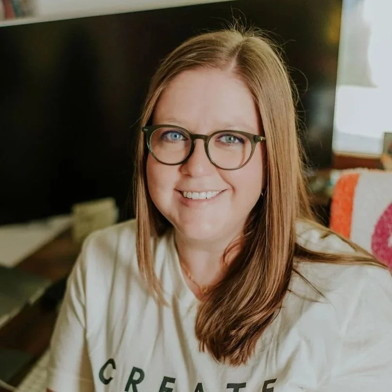 A woman with long red hair, glasses, and a beige shirt smiling at the camera in an indoor setting.