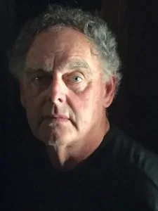 Close-up portrait of an older man with gray, curly hair and light skin, looking directly at the camera with a serious expression, against a dark background.