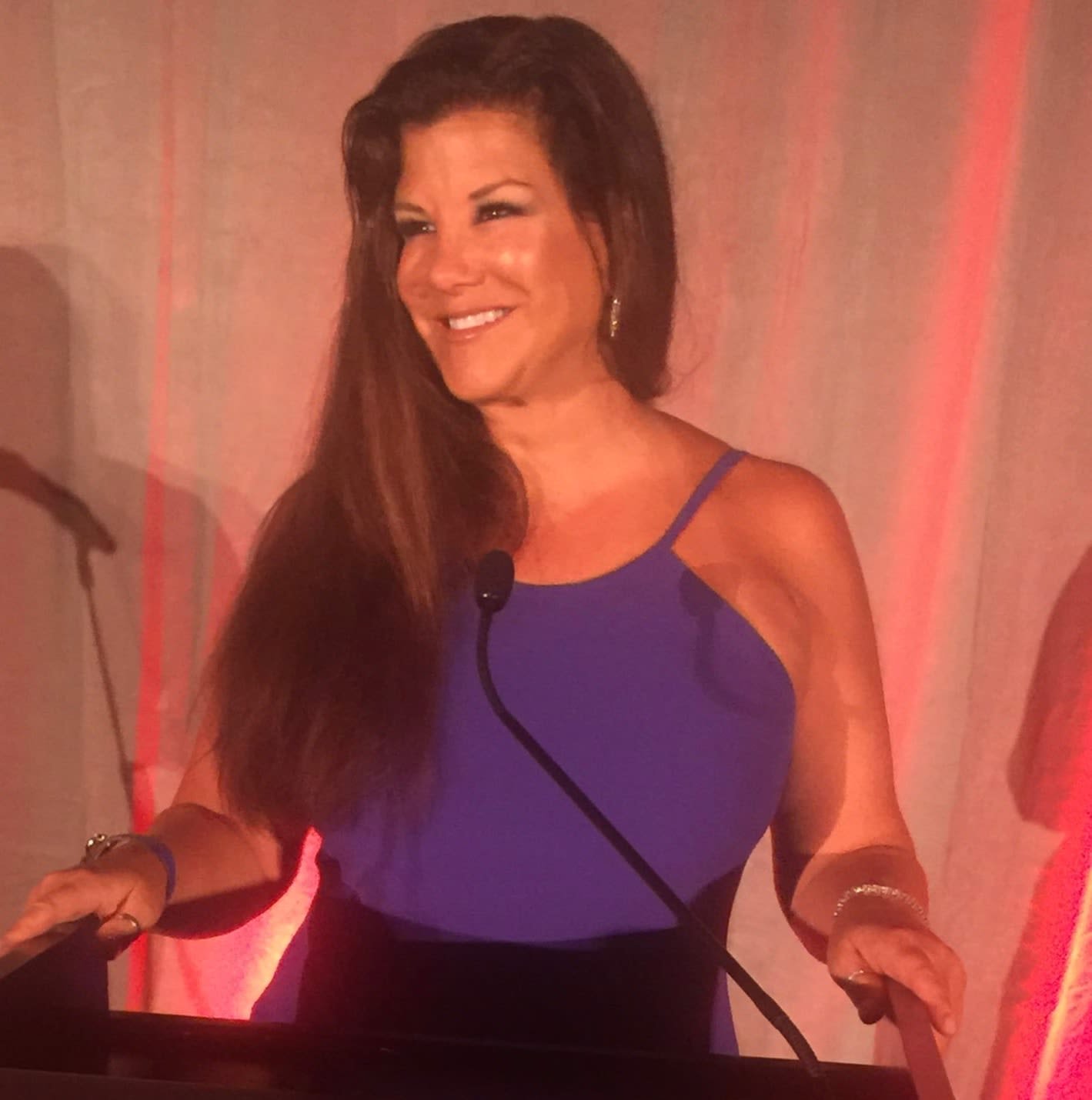 A woman with long dark hair wearing a blue dress, smiling and standing at a podium with a microphone.