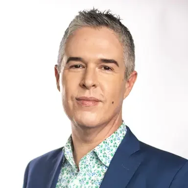 A man with short gray hair, wearing a blue suit jacket and a light green patterned shirt, looking serious against a plain white background.