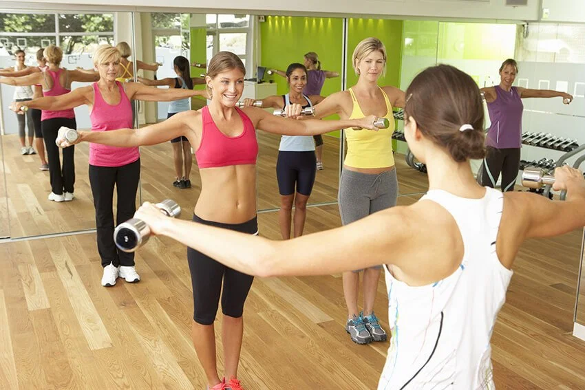 Small group personal training women performing a lateral arm raise exercise with dumbbells, facing an instructor in a mirrored fitness studio
