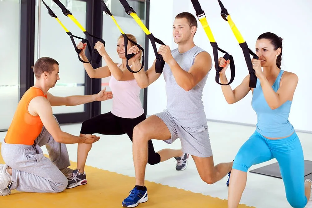 Male instructor shows men and women how to use TRX Training exquipment stretching down and pulling on the straps