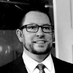 Black and white photo of a man wearing glasses and a suit, smiling.