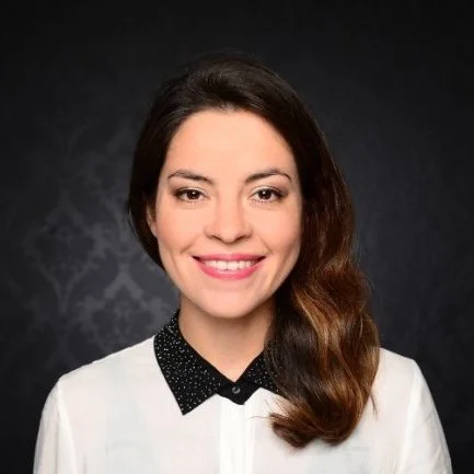 Portrait of a smiling woman with long brown hair, wearing a white blouse with a black collar, set against a dark, patterned background.