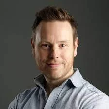 Portrait of a man with short hair, wearing a light blue shirt, smiling softly, with a neutral gray background.