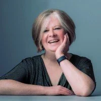 Smiling person with short, grey hair sitting at a table, wearing a dark top with hand resting on chin.