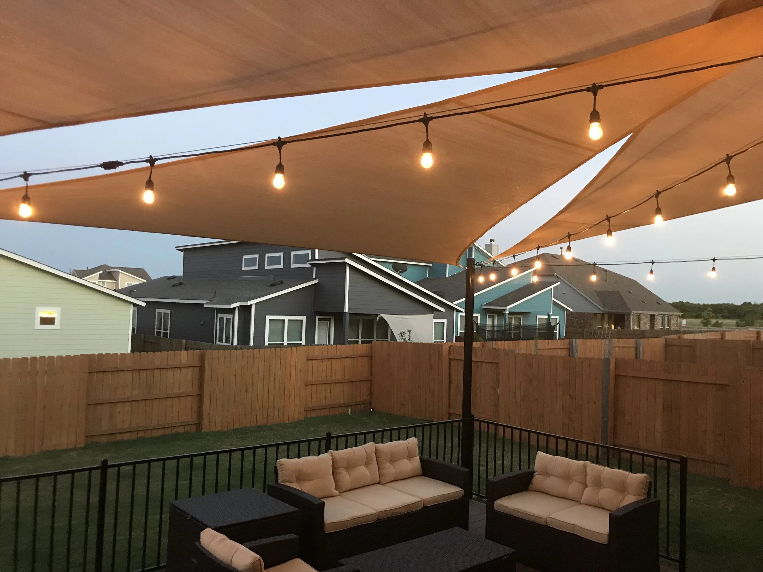 A backyard patio at dusk with beige cushioned outdoor furniture, including sofas and chairs, on a black metal frame. Overhead, large tan fabric sun shades are stretched between poles, with string lights hanging underneath. The backyard is enclosed by a wooden fence, and neighboring houses with dark and light siding are visible in the background.