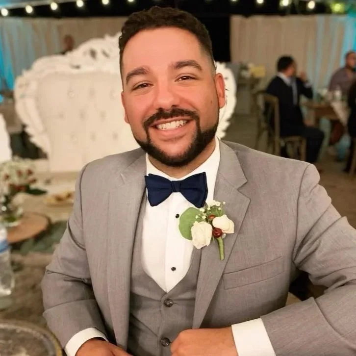 A man in a gray suit with a blue bow tie, smiling, at a formal event or wedding reception.