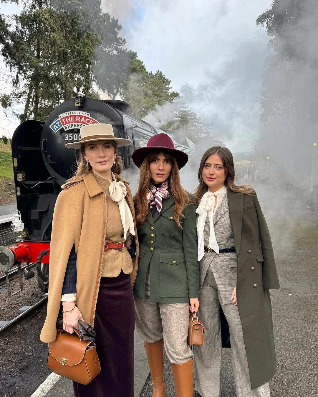 Three women dressed in vintage-style detective costumes posing in front of a black steam train with smoke coming out of its chimney, outdoors with trees in the background.