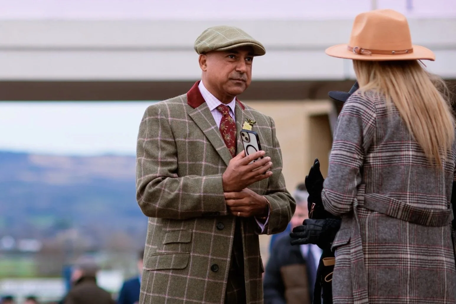 Man in a plaid suit holding a smartphone and talking to a woman wearing a plaid coat and tan hat at an outdoor event.