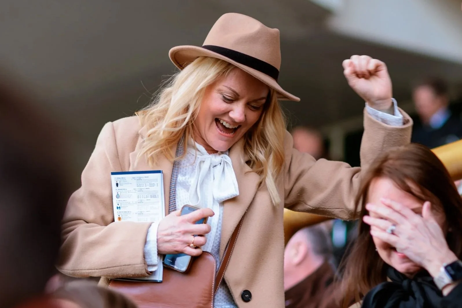 A woman in a beige coat and hat is celebrating with a fist pump while sitting next to another woman covering her face with her hand, in a crowded outdoor setting.