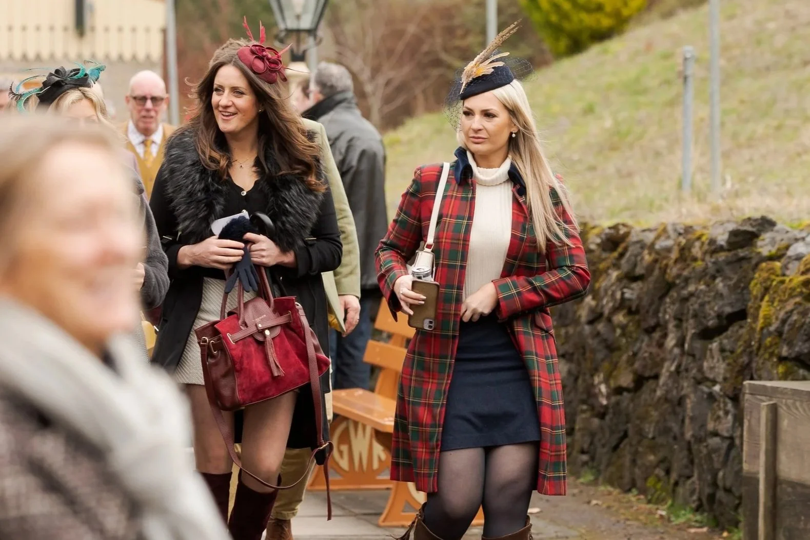 A group of women dressed in fashionable, winter clothing, some wearing hats and scarves, standing outdoors near a stone wall. The woman in the front is wearing a red tartan coat, black skirt, and sheer tights, holding a smartphone, with other women s