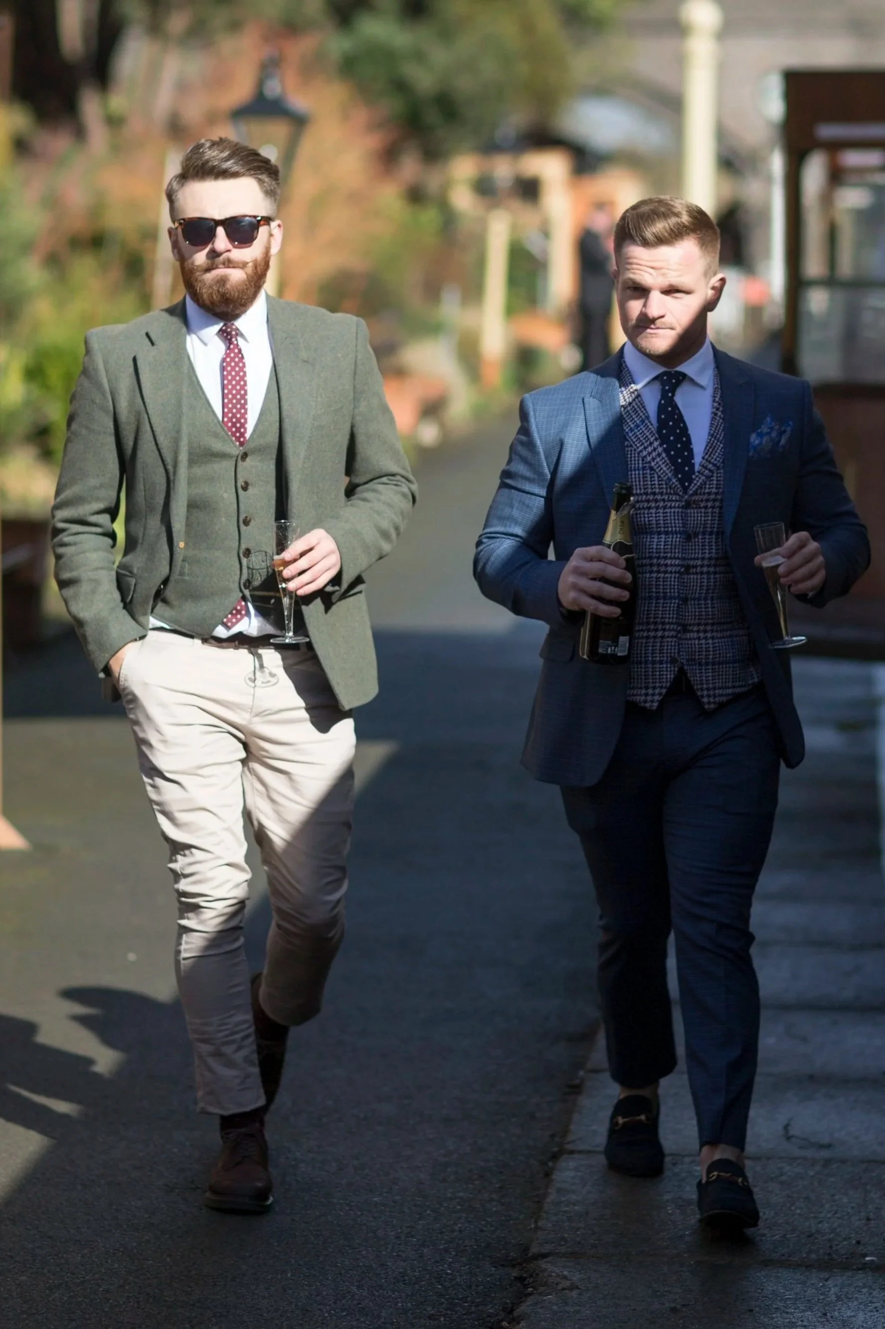 Two men in suits walking outdoors, each holding a champagne glass; one man with sunglasses and a beard, the other with short hair and a mustache. They are dressed stylishly, and the background shows a street with trees and buildings.