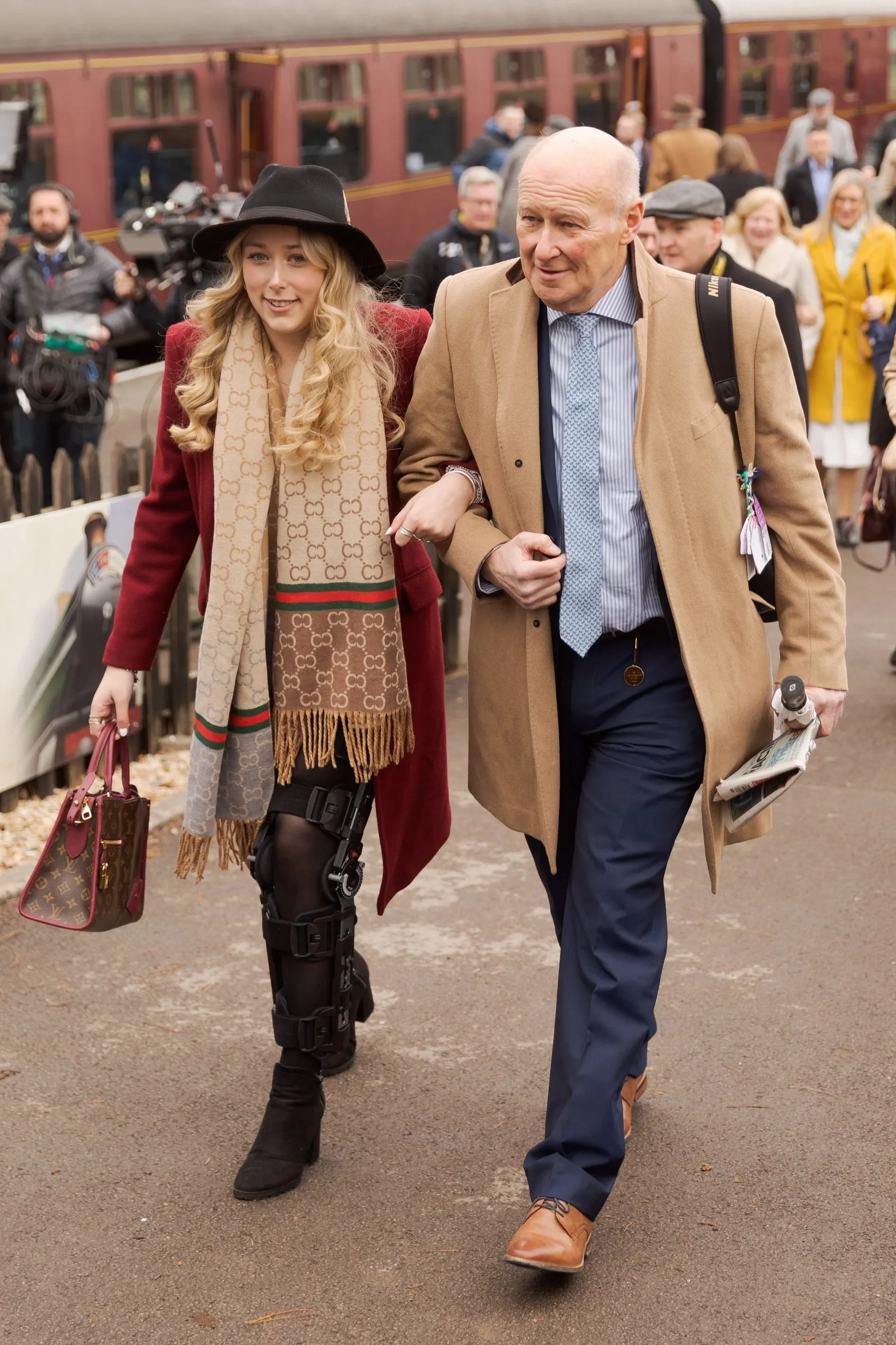 A young woman with long blonde hair, wearing a black hat, red coat, designer scarf, and a prosthetic leg, walking arm-in-arm with an older bald man in a tan coat, blue shirt, and dark trousers, on a train platform with a train and train station crowd