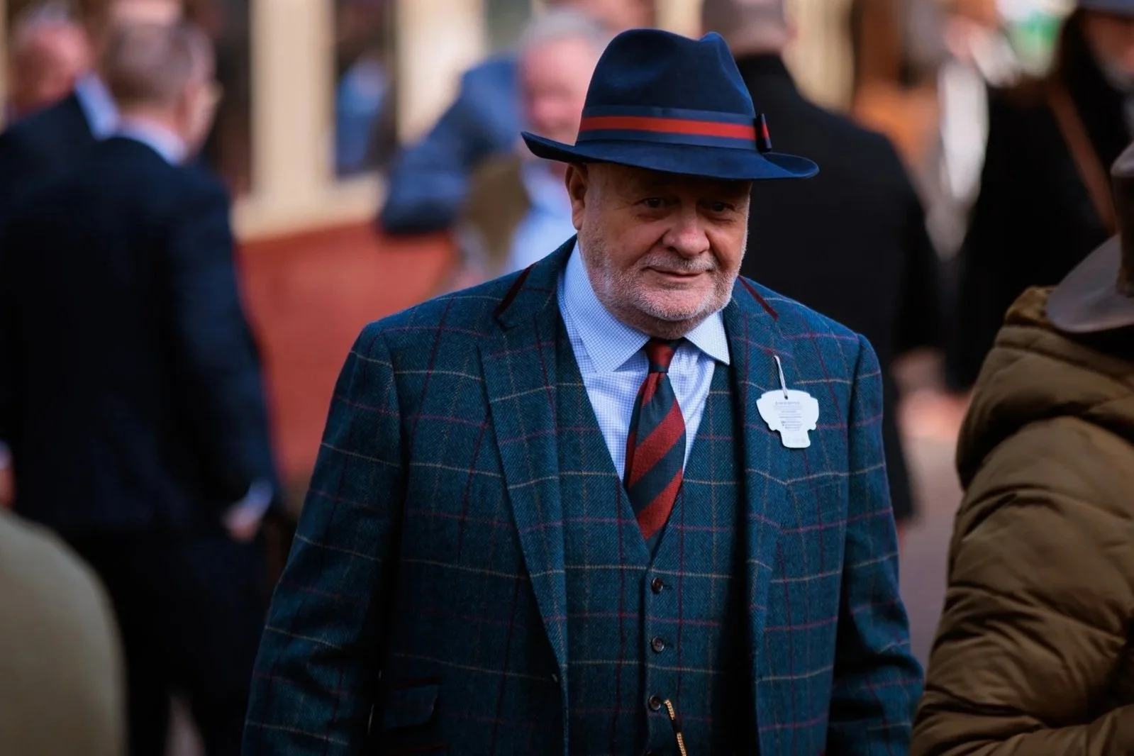 A man wearing a blue fedora hat and a plaid blazer walking in a crowded outdoor setting.