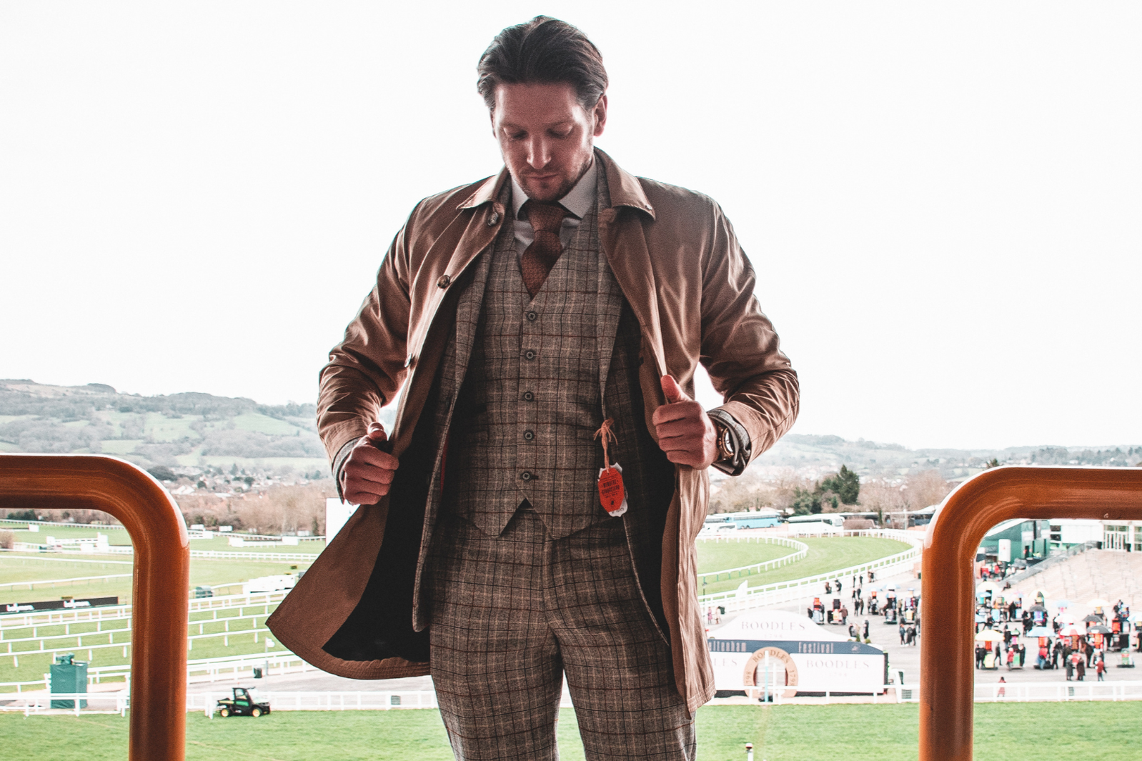 Man in a plaid suit and brown trench coat standing at the railing of a racetrack, with horses and people in the background.