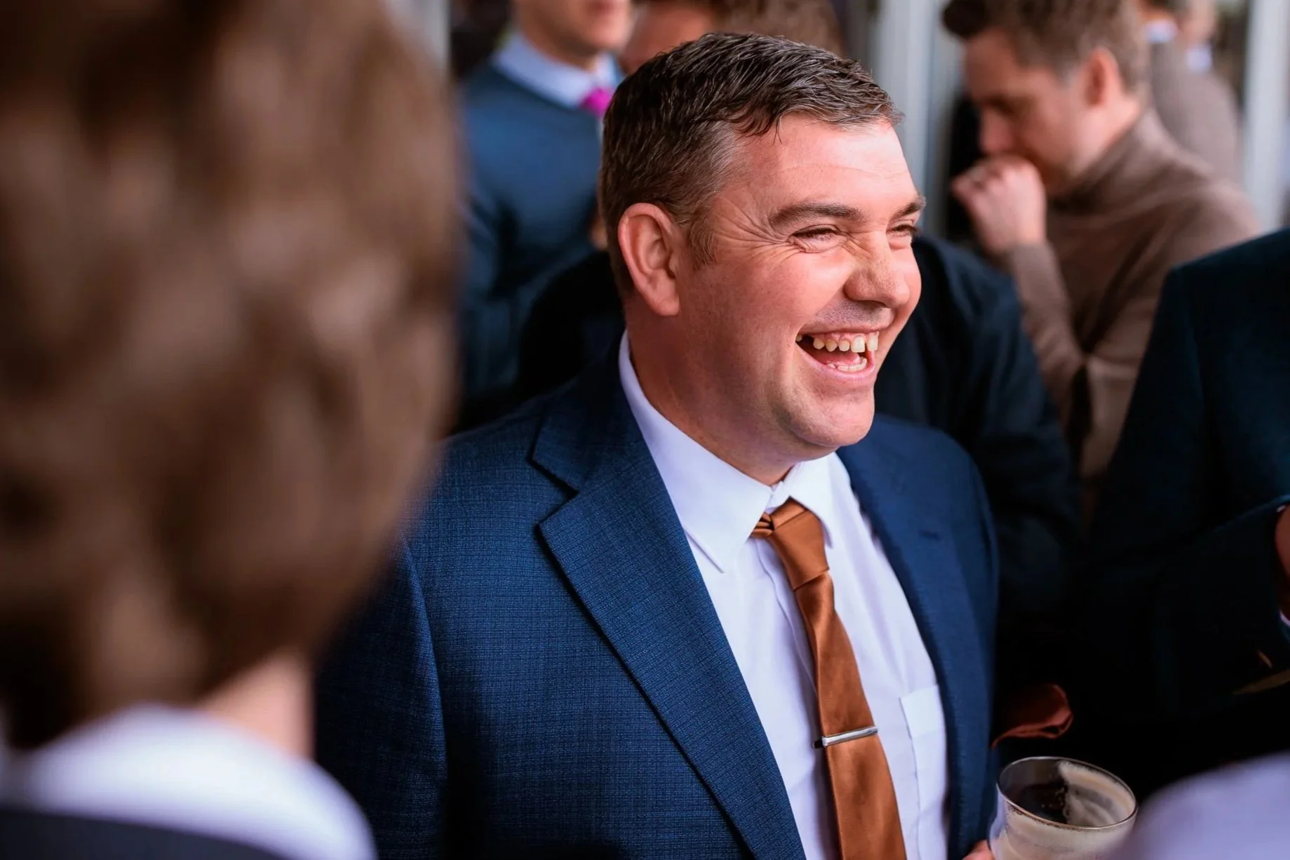 Man in a blue suit laughing and holding a drink at a social gathering or party