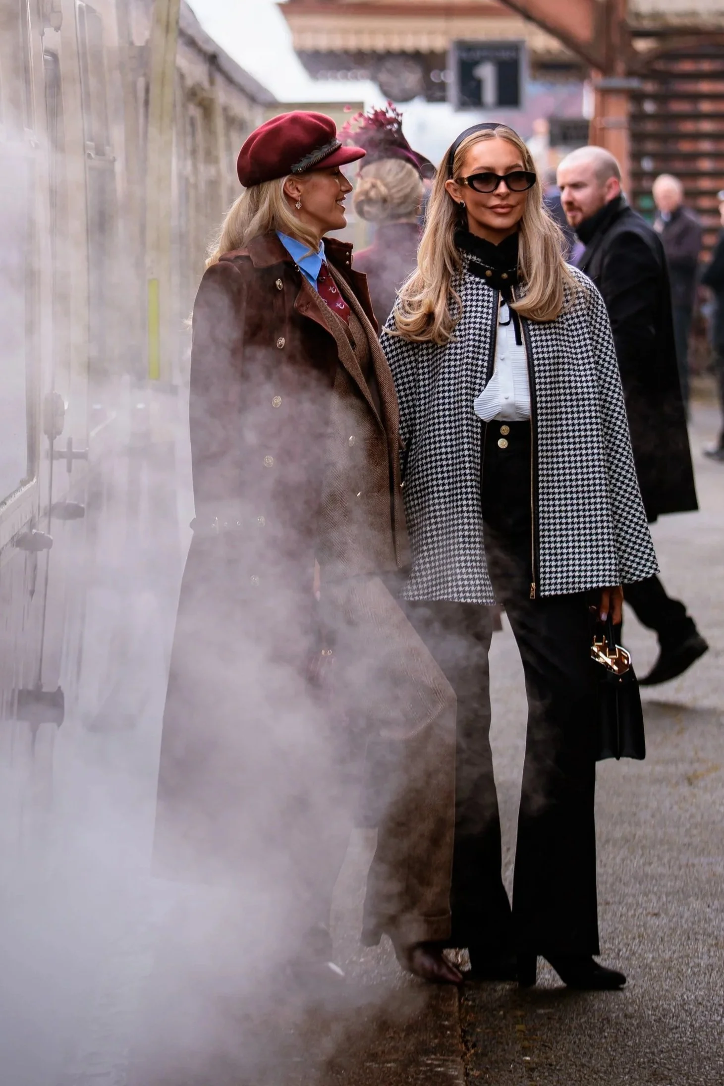 Two women standing outdoors near a steaming vent, dressed in stylish fall or winter clothing with coats and accessories.