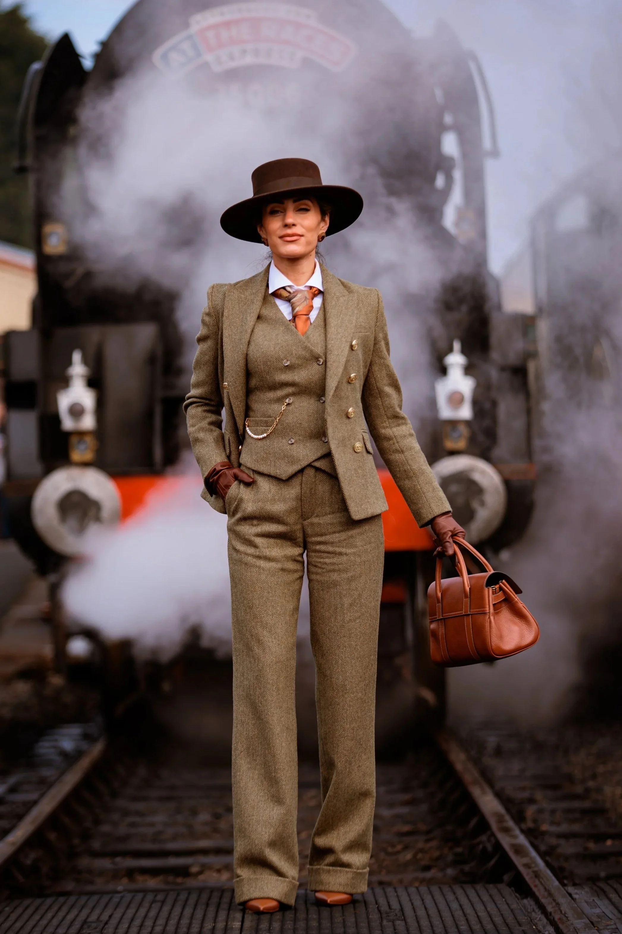 A woman dressed in vintage 1920s-style clothing standing on train tracks in front of a steam train, holding a brown leather handbag and wearing a wide-brimmed hat.